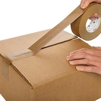 Person holding a roll of brown packing tape in their hand, with a cardboard box in the background