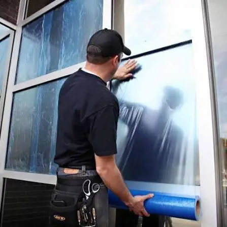 Man wearing a t-shirt, cap, and pants, holding a blue roll of window film in his hand