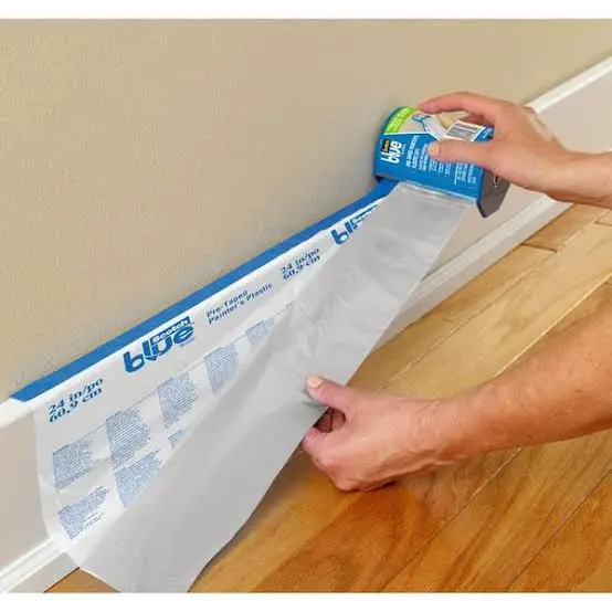 Person using a roll of blue tape to install a wall, with a paper on the wooden floor beneath them