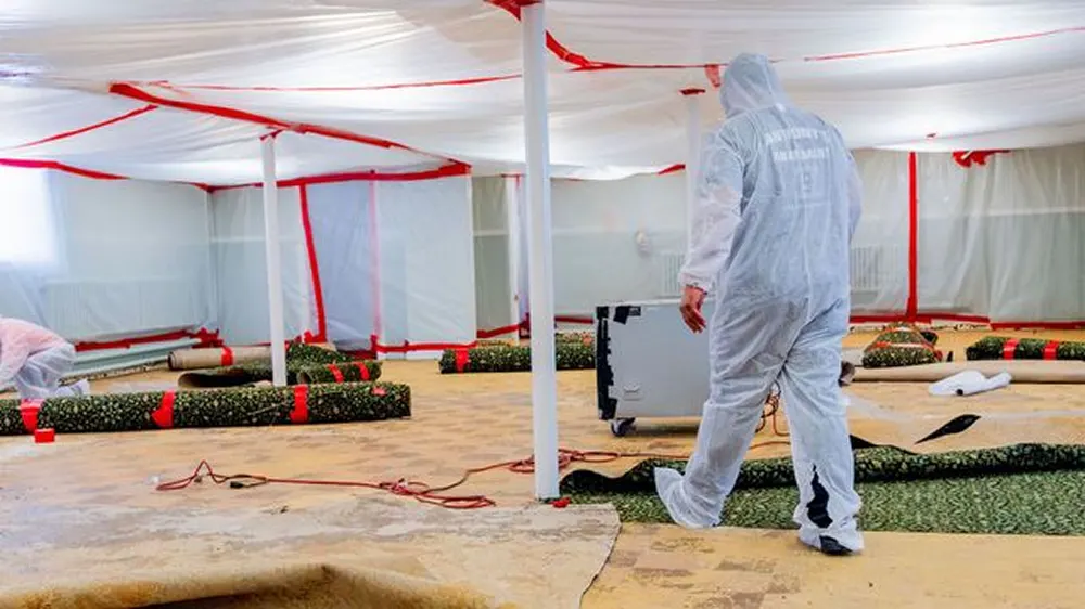 Man in a suit walking through a room filled with medical equipment, including a white tent with red ribbons